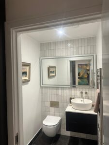 Property for sale by Auction This new home for sale features a modern bathroom with white tiled walls, a large mirror, and a rectangular sink on a sleek black vanity. A towel hangs from a rail beside it. A wall-mounted toilet sits beneath a framed picture, complementing the dark-colored floor—a Wimbledon gem indeed. Presented by World of Auctions