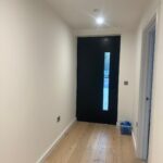 Property for sale by Auction A minimalist hallway in a Wimbledon new home for sale features light wooden flooring, white walls, and a black door with a vertical glass panel. A small blue box rests near the right wall, while ceiling lights warmly illuminate the space. Presented by World of Auctions