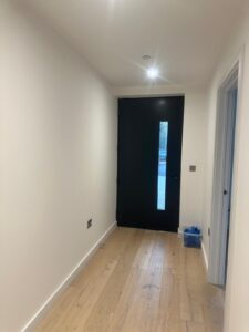Property for sale by Auction A minimalist hallway in a Wimbledon new home for sale features light wooden flooring, white walls, and a black door with a vertical glass panel. A small blue box rests near the right wall, while ceiling lights warmly illuminate the space. Presented by World of Auctions