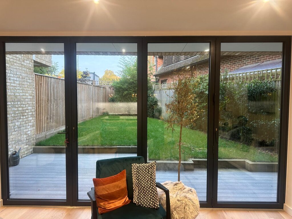 Property for sale by Auction Glass patio doors open to a backyard with a wooden deck and green lawn, offering serene views ideal for your new Wimbledon home. A chair with colorful cushions and a potted plant add to the inviting interior, while outside, a wooden fence and neighboring brick buildings provide charming neighborhood scenery. Presented by World of Auctions