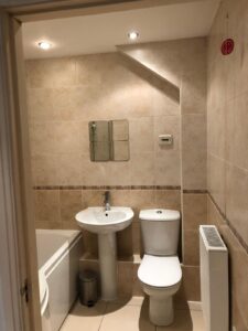 Property for sale by Auction A small bathroom with beige tiled walls features a white toilet, a pedestal sink with a small mirror above, and a curved bathtub. There is a radiator on the right wall and two ceiling lights. Presented by World of Auctions