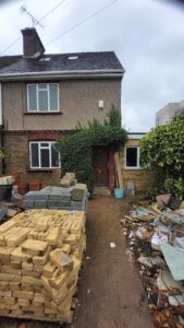 Property for sale by Auction A partially renovated house with a worn exterior. Bricks and construction materials are scattered in the front yard. A small door is partially covered by climbing plants, and a side extension is visible. Overcast sky above. Presented by World of Auctions