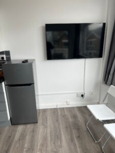 Property for sale by Auction Located in London NW6, this small, modern 1-bed flat offers an enticing investment opportunity. It features a wall-mounted flat-screen TV, a compact gray refrigerator with a remote control on top, and two white folding chairs on a laminate wood floor. Gray-curtained windows complete the chic look. Presented by World of Auctions