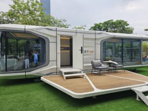 Property for sale by Auction A modern, modular home with large windows and sleek design sits on a grassy area. It has an open door leading to the interior, and a small deck with two chairs in front. Trees and a building are visible in the background. Presented by World of Auctions