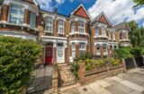 Property for sale by Auction A row of charming Victorian terraced houses with red brick facades and ornate gables. The front gardens are enclosed by low brick walls, featuring lush greenery and flowers. A clear blue sky with a few clouds is visible above. Presented by World of Auctions