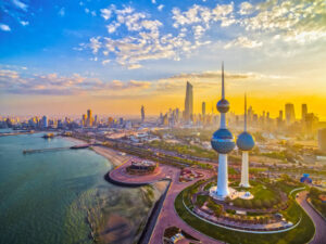 Property for sale by Auction A stunning aerial view of Kuwait City at sunset, featuring the iconic Kuwait Towers. The sky is dotted with clouds, and the cityscape is illuminated by the golden glow of the setting sun, with the sea visible to the left. Presented by World of Auctions