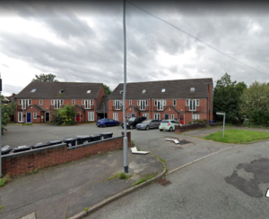 Property for sale by Auction A suburban street scene with a block of brick townhouses for sale. Cars are parked in the driveways, and a row of trash bins is visible on the left. The sky is overcast, and trees are in the background, providing a quaint touch to this Newcastle Stoke on Trent neighborhood. Presented by World of Auctions