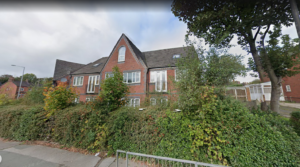 Property for sale by Auction A two-story brick building with a triangular roof, part of a block of investment apartments for sale, is partially obscured by tall trees and bushes. The cloudy sky looms over a small street and fence in the foreground, nestled in picturesque Newcastle Stoke on Trent. Presented by World of Auctions