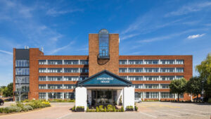 Property for sale by Auction A modern, brick office building under a clear blue sky, labeled "Arrowhead House" above the entrance. The facade features a symmetrical design with five stories and large, reflective windows. A small landscaped area is in front. Presented by World of Auctions