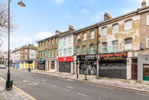 Property for sale by Auction A deserted urban street with a row of old brick buildings. Shops on the ground floor have closed shutters. The road is empty, and there's a bus stop sign on the sidewalk. The sky is clear. Presented by World of Auctions