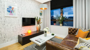 Property for sale by Auction A modern living room features a brown leather sofa with yellow and patterned cushions. A large window is adorned with curtains. The room includes a wall-mounted TV, decorative vases with flowers, a white coffee table, and framed artwork on a textured wall. Presented by World of Auctions