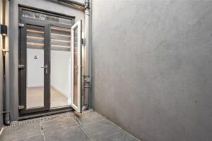 Property for sale by Auction A small, enclosed outdoor space with gray walls and a tiled floor. There's a glass door partially open, revealing a glimpse of an interior room with light-colored walls and a wooden floor. The area appears modern and minimalistic. Presented by World of Auctions