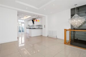 Property for sale by Auction Spacious modern interior with a white kitchen, featuring sleek cabinetry, pendant lights, and a tiled floor. A chandelier hangs above the open dining area with glass balustrade and polished tile flooring, creating an elegant atmosphere. Presented by World of Auctions