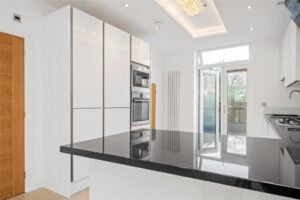 Property for sale by Auction Modern kitchen with white cabinets, built-in appliances, and a glossy black countertop island. The room features a wooden door, a large window, and recessed ceiling lights, creating a bright and open space. Presented by World of Auctions