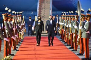 Property for sale by Auction Two formally dressed individuals walk down a red carpet, flanked by a military honor guard in uniforms with plumed hats and rifles. A staircase from an aircraft is visible in the background. Presented by World of Auctions