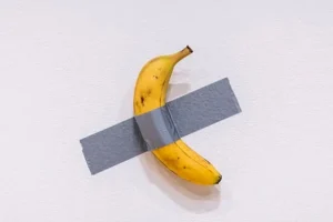 Property for sale by Auction A banana is taped to a white wall with a piece of gray duct tape. The banana is positioned diagonally with a slight curve. The wall is plain and untextured, creating a minimalist background for the artwork. Presented by World of Auctions