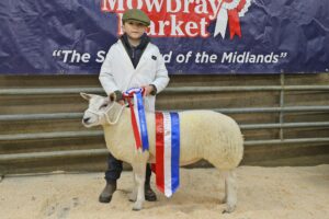 Property for sale by Auction A young person wearing a cap and white coat stands beside a sheep adorned with a red, white, and blue award ribbon. They are in front of a banner reading "Melton Mowbray Market". The scene appears to be at an animals' market or competition. Presented by World of Auctions