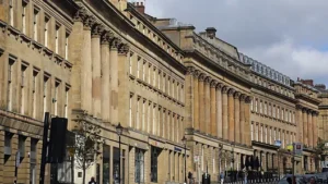 Property for sale by Auction Street view of a historic city with rows of Georgian-style stone buildings featuring large windows and ornate columns. The cloudy sky is overhead, and pedestrians and cars are visible on the road. Presented by World of Auctions