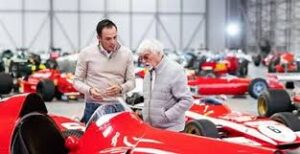 Property for sale by Auction Two men stand near a red vintage race car in a large garage filled with classic cars. One man wears a beige sweater; the other is in a gray jacket. They appear to be engaged in conversation, with numerous cars visible in the background. Presented by World of Auctions