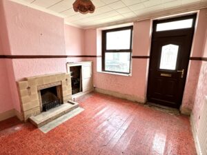 Property for sale by Auction A small empty room with pink textured walls and a red patterned floor is featured in this investment property for sale on Padley Hill, Mansfield. It includes a fireplace with a beige mantel, two large windows, a dark brown front door, and a hanging light fixture on the ceiling. Presented by World of Auctions