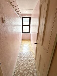 Property for sale by Auction A narrow hallway with pink textured walls and a light bulb dangling from the ceiling leads to a wooden door slightly ajar. The patterned linoleum floor and hooks on the left wall add character, while natural light streams through a window at the end. Perfect investment property for sale in Padley Hill, Mansfield NG18. Presented by World of Auctions
