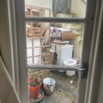 Property for sale by Auction View through a window of a cluttered outdoor area, typical of an investment apartment in Brighton BN3 for sale by auction. A potted plant, washing machine, chair, and various items rest on weathered concrete beside a tall fence and wooden lattice. Presented by World of Auctions