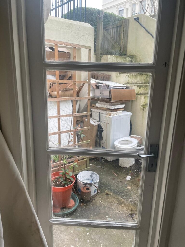 Property for sale by Auction View through a window of a cluttered outdoor area, typical of an investment apartment in Brighton BN3 for sale by auction. A potted plant, washing machine, chair, and various items rest on weathered concrete beside a tall fence and wooden lattice. Presented by World of Auctions
