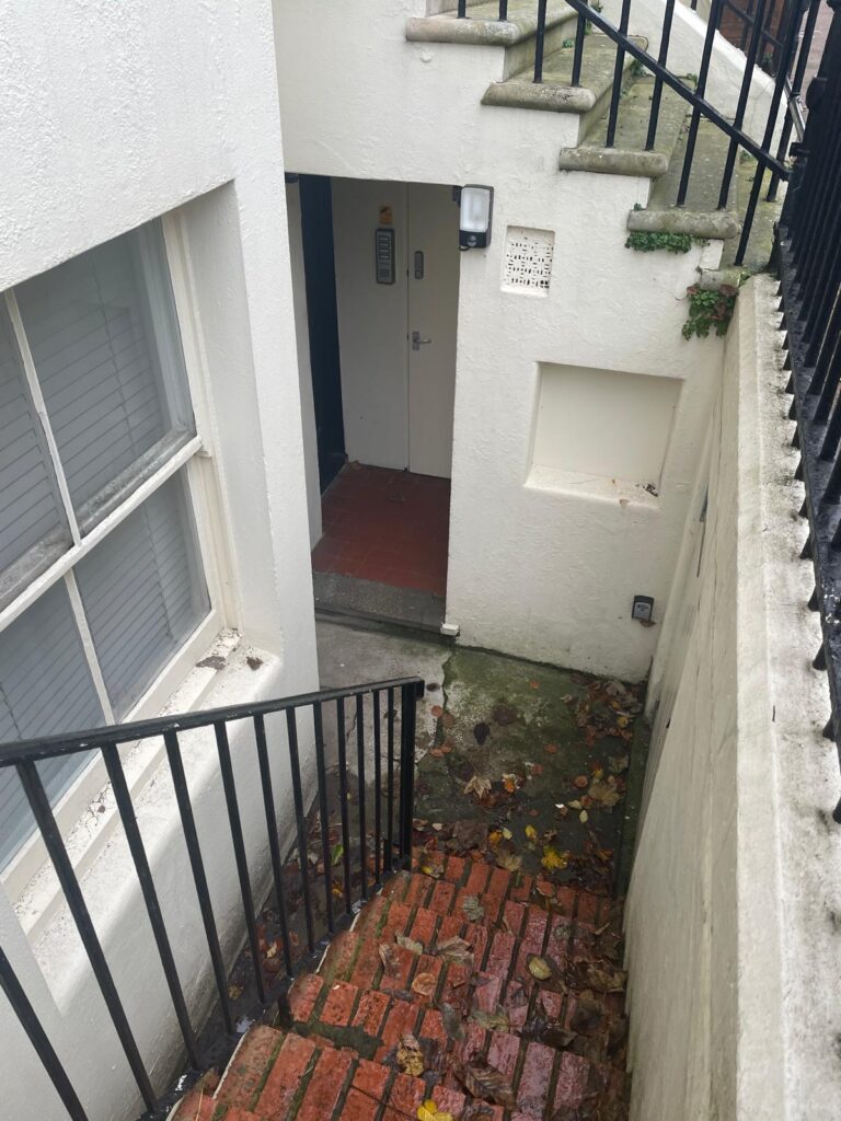 Property for sale by Auction A narrow staircase with black handrails guides you down to the basement entrance of this charming investment apartment. The red brick steps, dotted with leaves, lead to a pristine white door and walls, accented by a small window in Brighton BN3, for sale by auction. Presented by World of Auctions