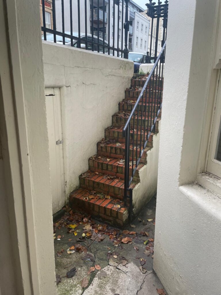 Property for sale by Auction A narrow, outdoor brick staircase with a black metal railing ascends from the lower level, embraced by white walls. Fallen leaves add charm to the steps of this investment apartment in Brighton BN3, set against a backdrop of buildings and up for sale by auction. Presented by World of Auctions