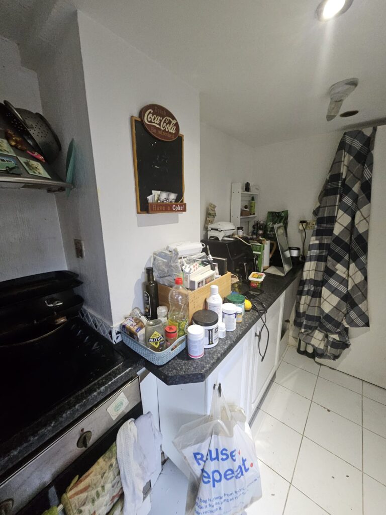 Property for sale by Auction The cluttered kitchen counter in this charming investment apartment in Brighton BN3 for sale by auction features various food items, containers, and appliances. A Coca-Cola mirror hangs above, while a checkered cloth is draped on the right. The white floor contrasts with dark countertops. Presented by World of Auctions