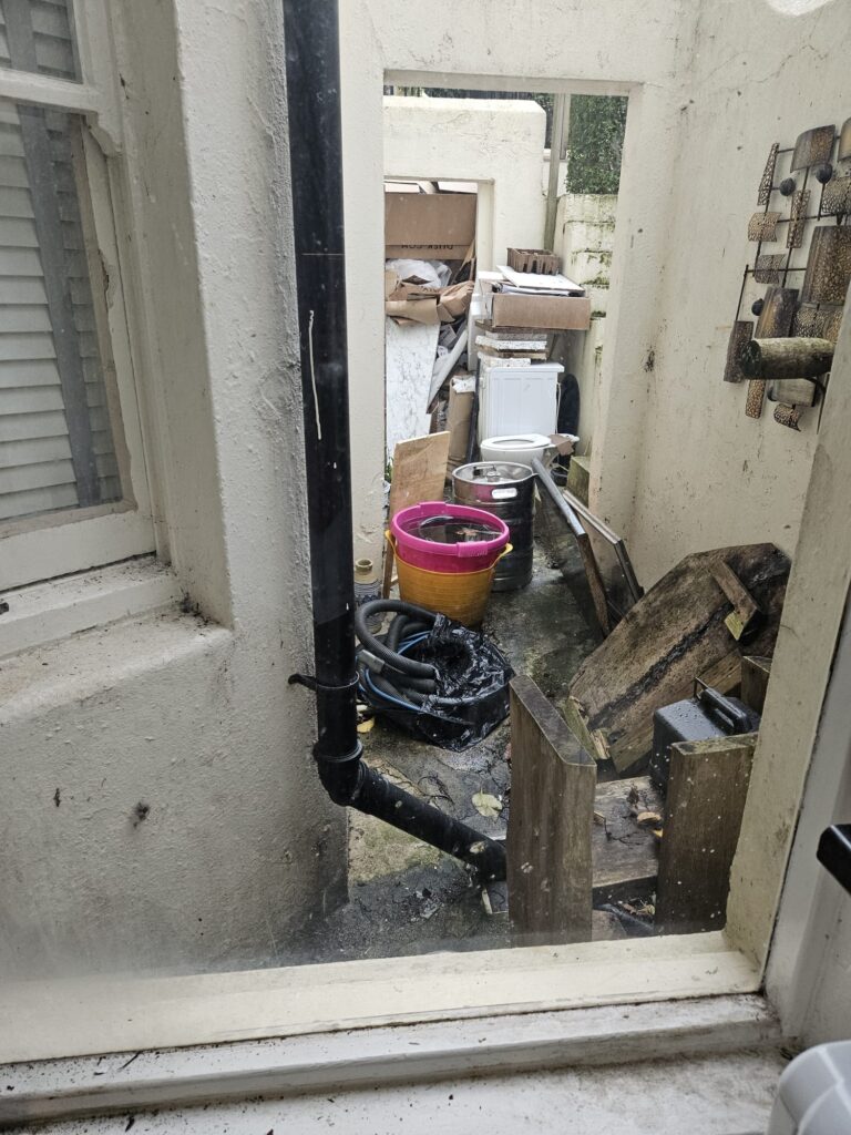 Property for sale by Auction A cluttered outdoor area, visible through a window, hints at potential in this investment apartment in Brighton BN3 for sale by auction. With items like buckets, hoses, and a washing machine scattered about, the space appears narrow and messy, featuring a drainpipe and window on the left. Presented by World of Auctions