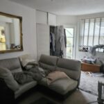 Property for sale by Auction This Brighton BN3 investment apartment, available for sale by auction, features a cozy living room with a gray sofa, pillows, and a blanket. A large mirror adorns the wall beside an open wardrobe. Natural light pours through vertical blinds, highlighting an exercise machine and cozy clutter in the corner. Presented by World of Auctions